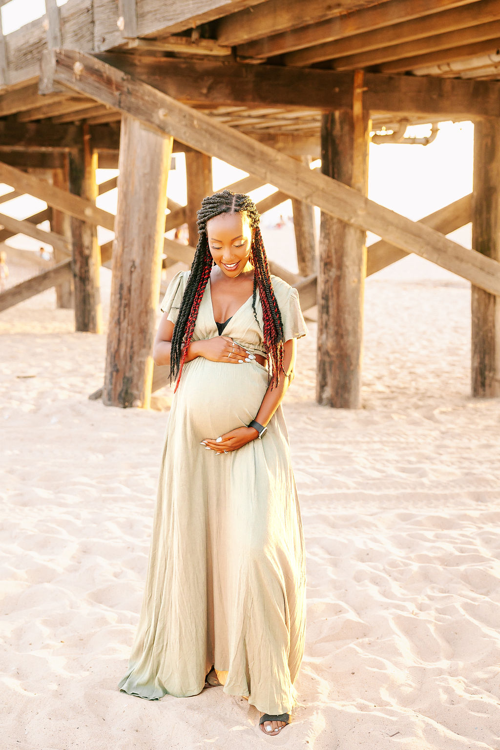 A happy mom to be walks on the beach under a wooden pier at sunset in a gold maternity gown smiling down at her bump after some prenatal massage in boise