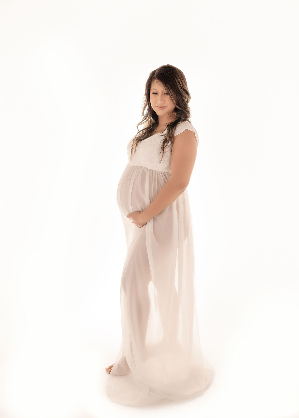 A pregnant woman in a white maternity gown smiles while gazing down at her bump in her hands after some prenatal yoga in Boise