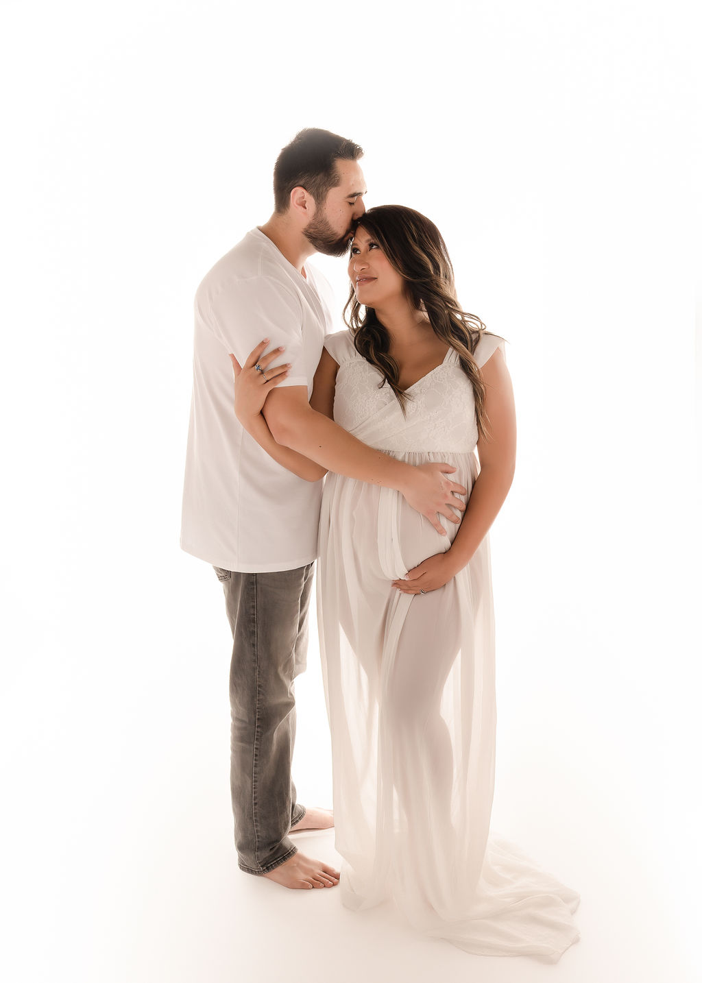 A happy expecting couple snuggles while kissing the mom's head in a studio in a white maternity gown after some prenatal yoga in Boise